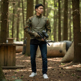 id-paintball Combat Shirt in Flecktarn mit elastischem Torso und Rip-Stop-Ärmeln, getragen von einem Mann im Wald.