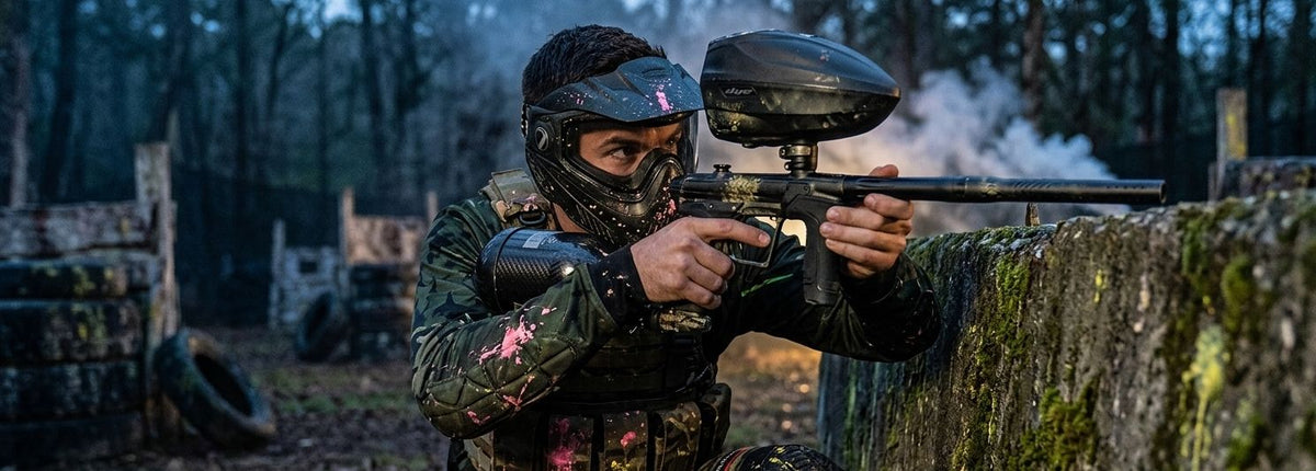 Paintballspieler mit Maske und Markierer beim Woodland-Paintball hinter einer Deckung.