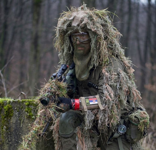 Paintballspieler im Ghillie-Anzug mit getarntem Markierer im Wald.